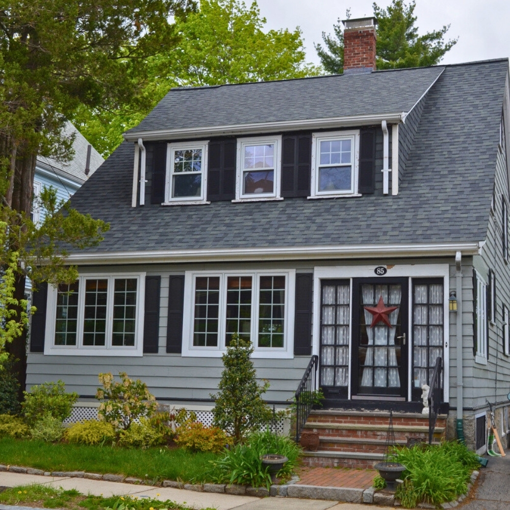 New roof on a small two-story house in Massachusetts with gray siding and black shutters. Trees surrounding.