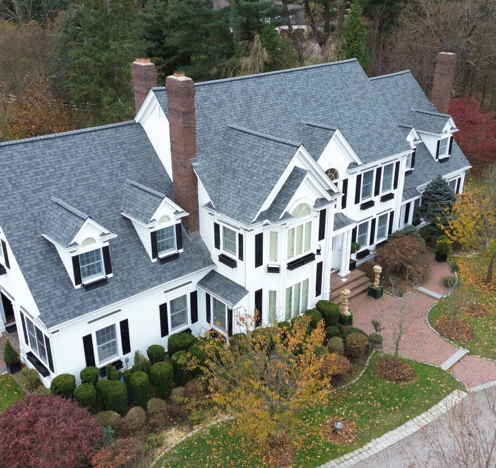 Angled upper view of large white two-story house with black shutters, multiple dormers, and newly installed gray roof. 