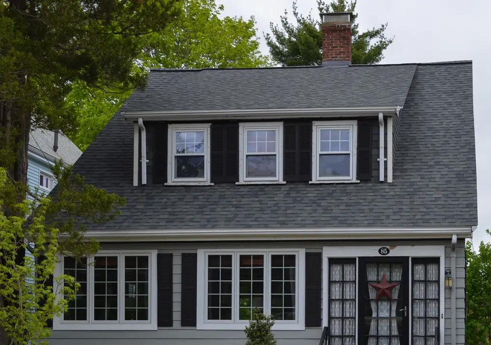 Front view of small two-story gray house with black shutters and a new gray-shingled roof, three-window dormer, trees surrounding.