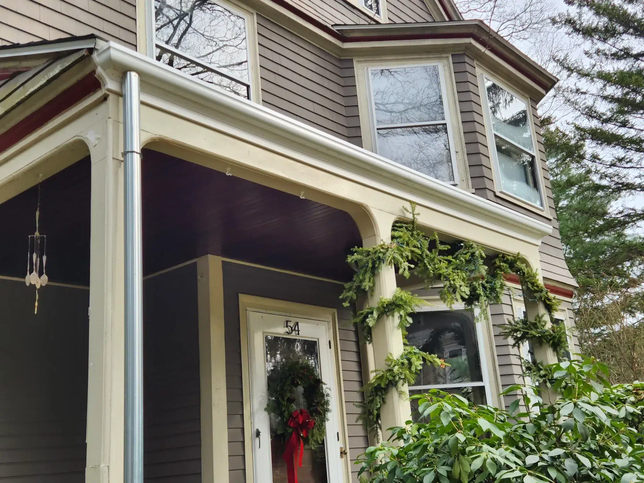Fiberglass gutters newly installed on a Victorian style home with brown siding and off-white trim. Wreath on front door and pine garland wrapped around front porch columns.