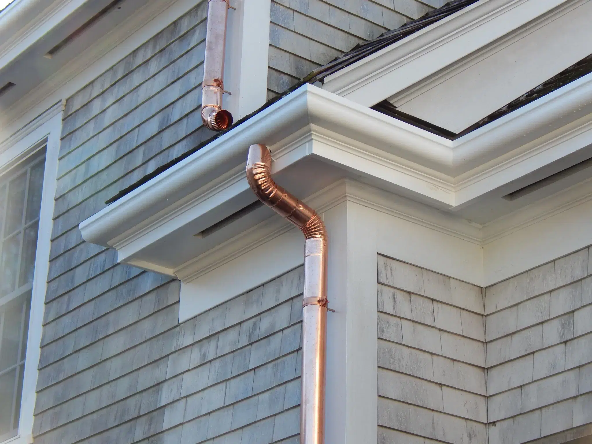 Fiberglass gutters with copper downspout installed on the side of a two-story home with light gray cedar siding.